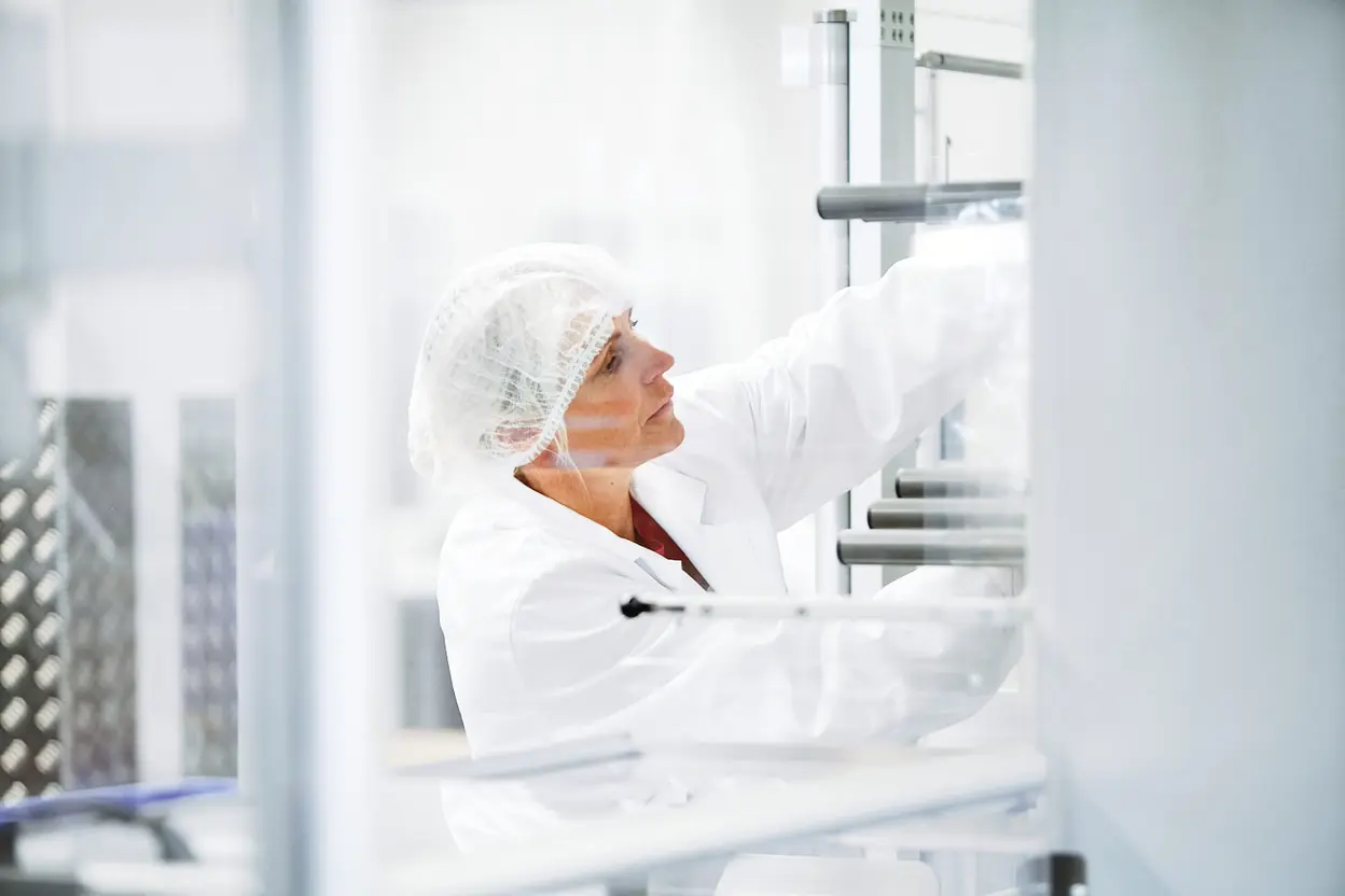 Mitarbeiterin in weißem Laborkittel und Haarnetz stellt etwas an einer Produktionsanlage ein.