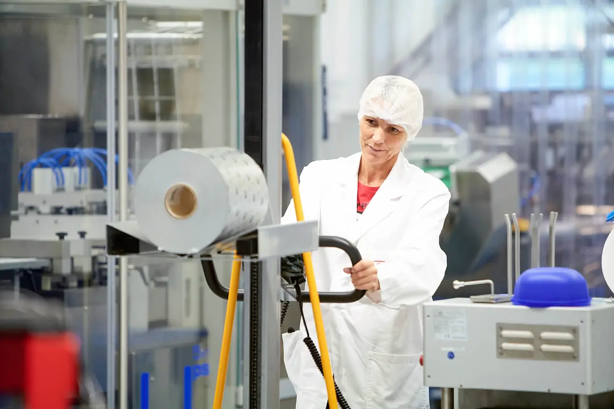 Mitarbeiterin in weißem Laborkittel und Haarnetz bedient einen Hubwagen neben einer Verpackungsmaschine mit großer Kunststoff-Folienrolle.