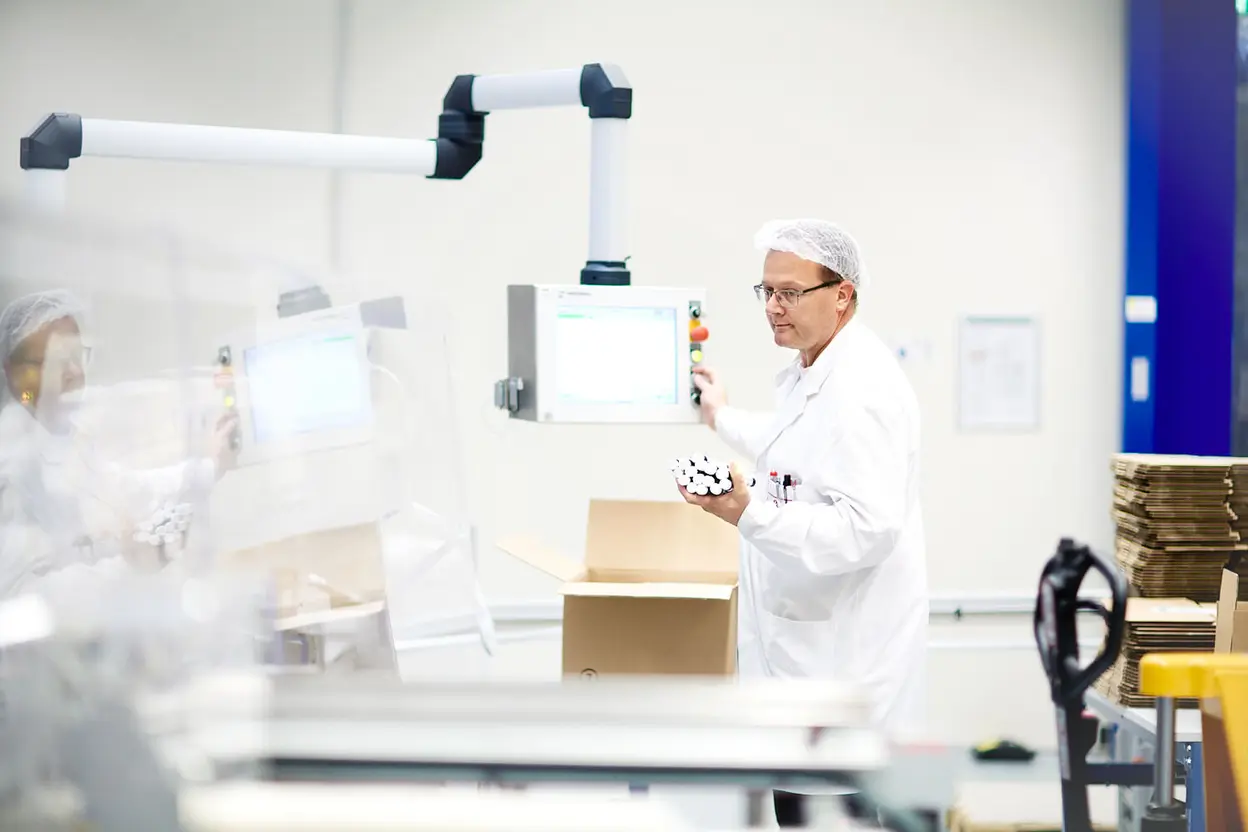 Mitarbeiter im weißen Laborkittel und Haarnetz steht an einer Verpackungslinie und bedient ein Bedienpanel.