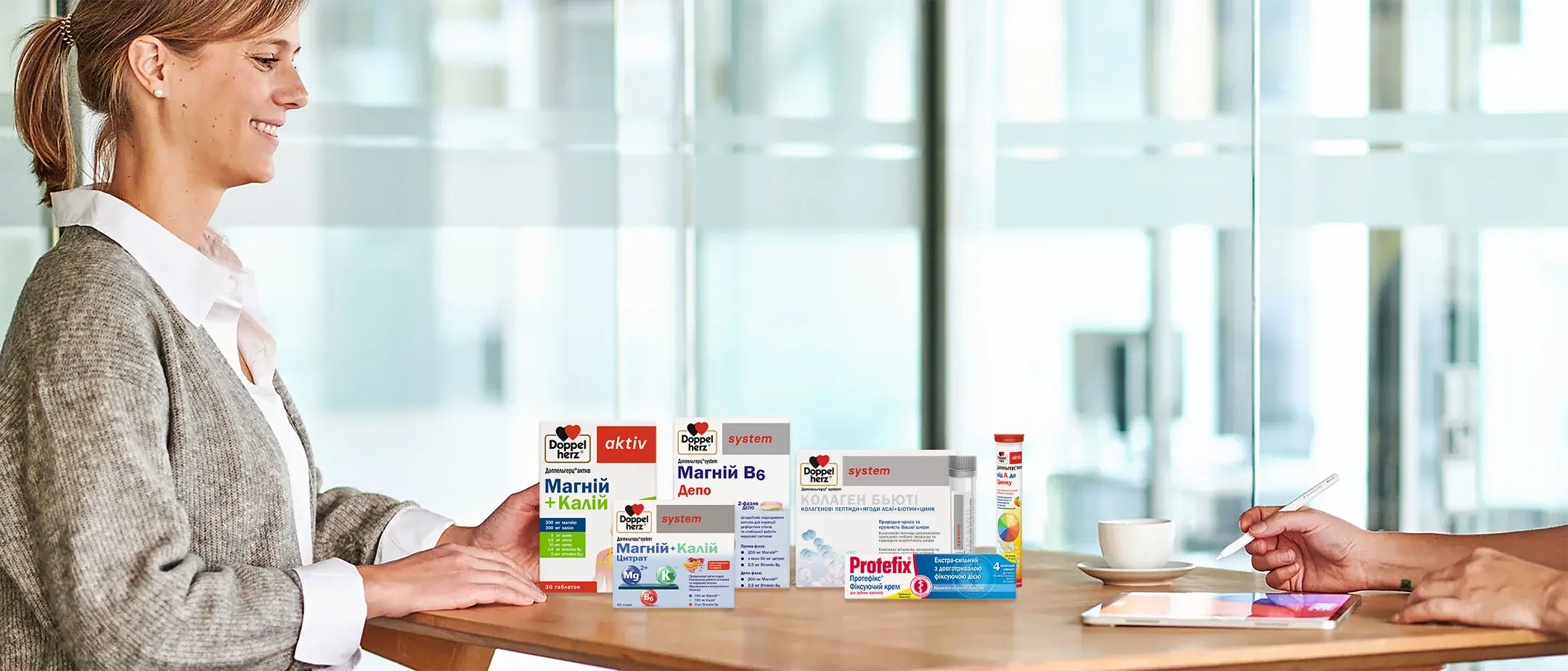 A woman sits at a table, smiling, while facing a selection of health product boxes displayed prominently. A cup and a digital tablet with a stylus are also present on the table, suggesting a consultation about the products.