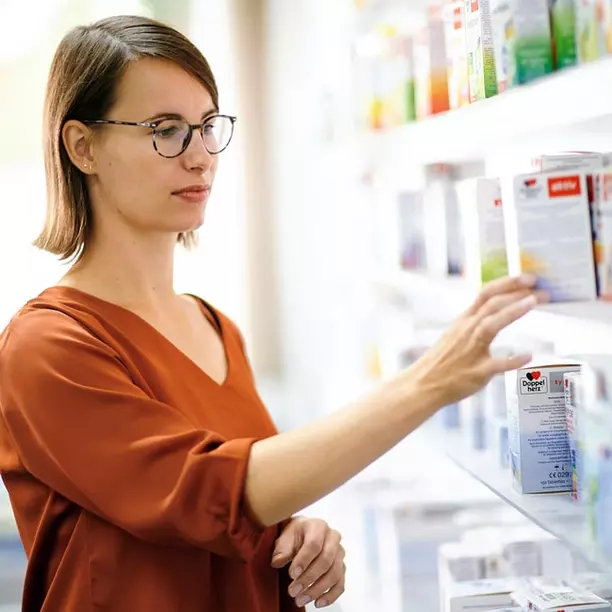 A woman with short hair and glasses is reaching for a product on a store shelf lined with colorful boxes. She is wearing an orange blouse and appears focused on her selection. The background is bright and slightly blurred.