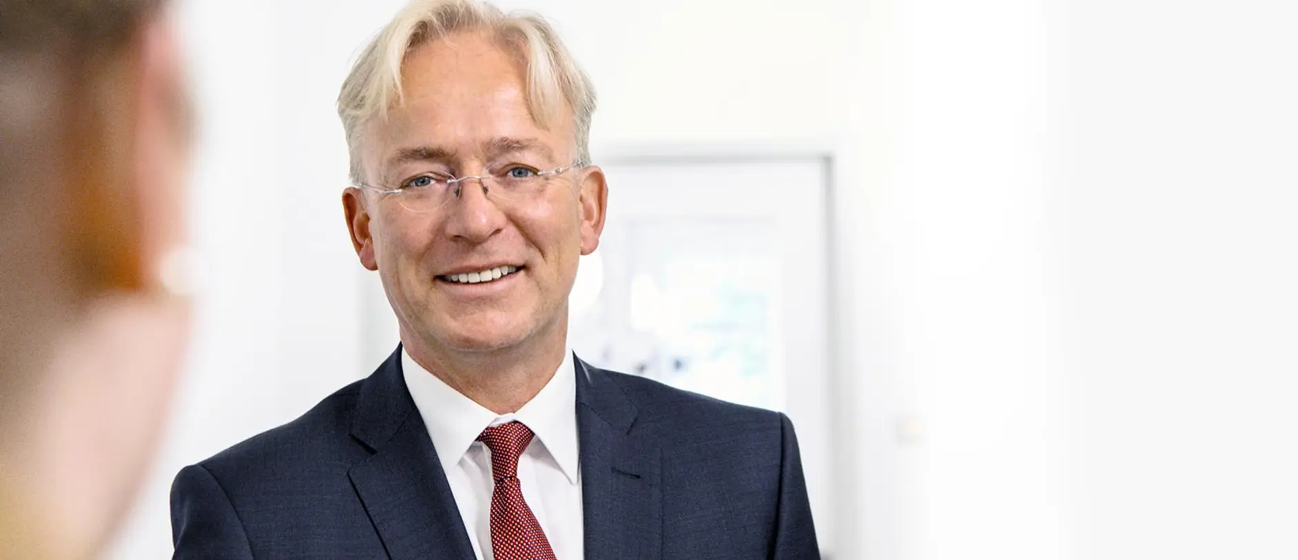 A smiling man with glasses and light blonde hair is wearing a dark suit and a red tie. He appears to be engaged in a conversation and is positioned against a blurred light-colored background. The setting suggests a professional environment.