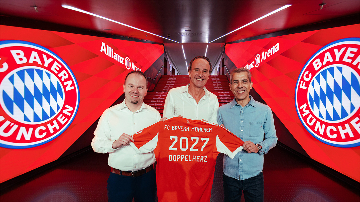 Троє чоловіків стоять на фоні червоного освітлення в Allianz Arena. Один з них тримає футболку FC Bayern München з написом "2027" та "Doppelherz". Всі виглядають усміхненими і святковими.