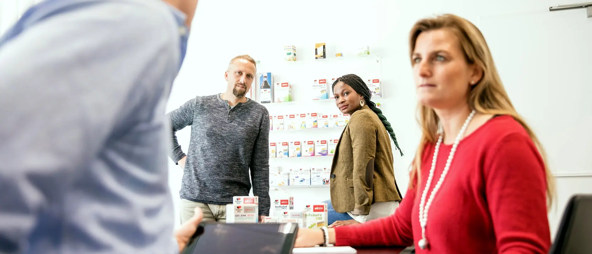 A diverse group of three professionals is engaged in a discussion. One person stands in the background, while two others are seated at a table with a digital device in front of them. Behind them is a display of various products on shelves.