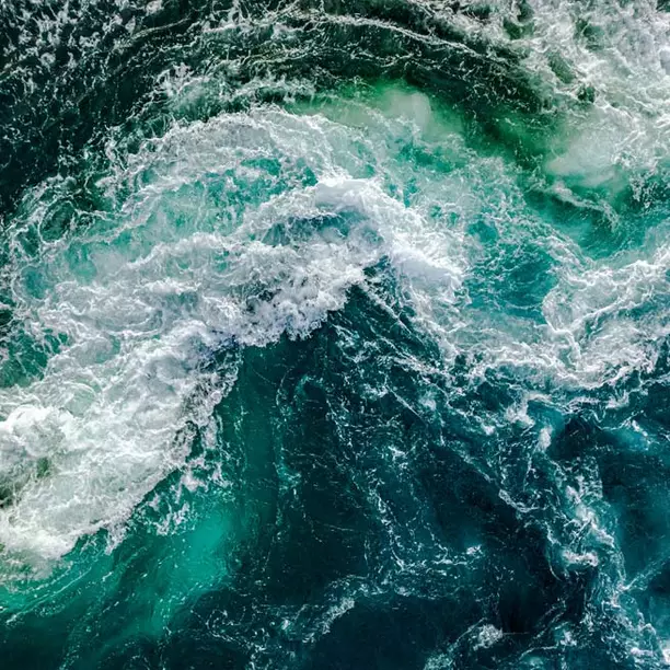 An aerial view of ocean waves creating a dynamic pattern. The water shows deep blue and turquoise hues, with white foam and swirling currents, reflecting a vibrant and lively marine environment.