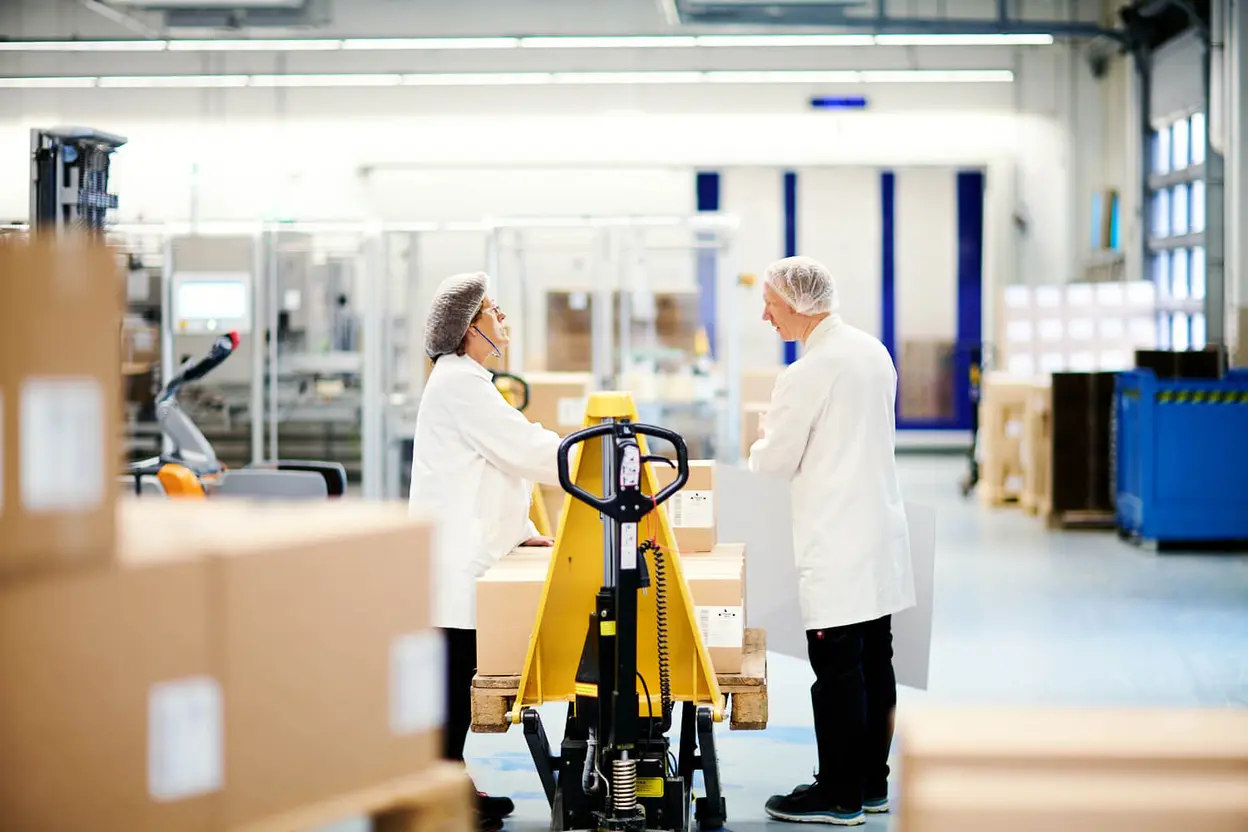Zwei Mitarbeitende in weißem Laborkittel und Haarnetz stehen in einer Verpackungshalle an einem Hubwagen und unterhalten sich zwischen Kartons.