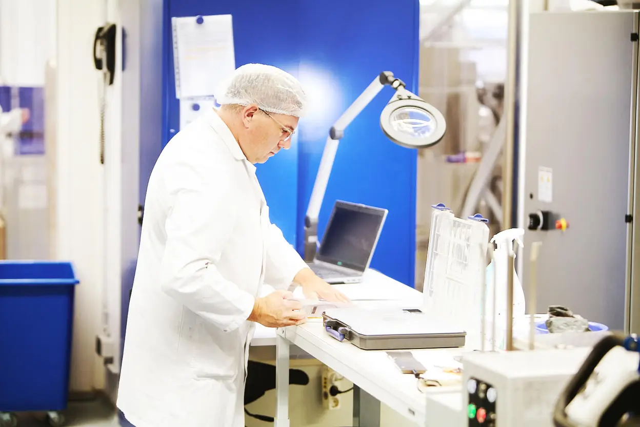 Mitarbeiter mit Haarnetz und Laborkittel arbeitet an einem Labortisch mit einem geöffnete Koffer und einem Laptop, im Hintergrund eine blaue Trennwand.