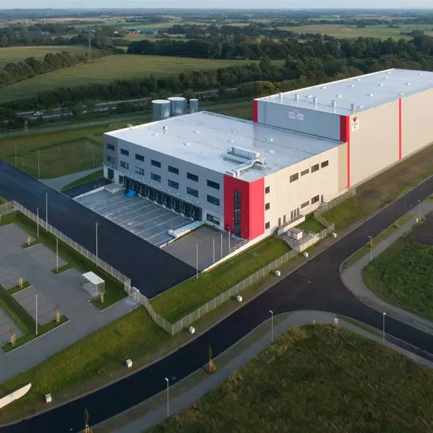 A large, modern warehouse building is depicted, featuring a gray exterior with a prominent red section. Loading bays are visible in the front, surrounded by paved roads and green fields in the background. The structure is well-lit, indicating an operational facility.